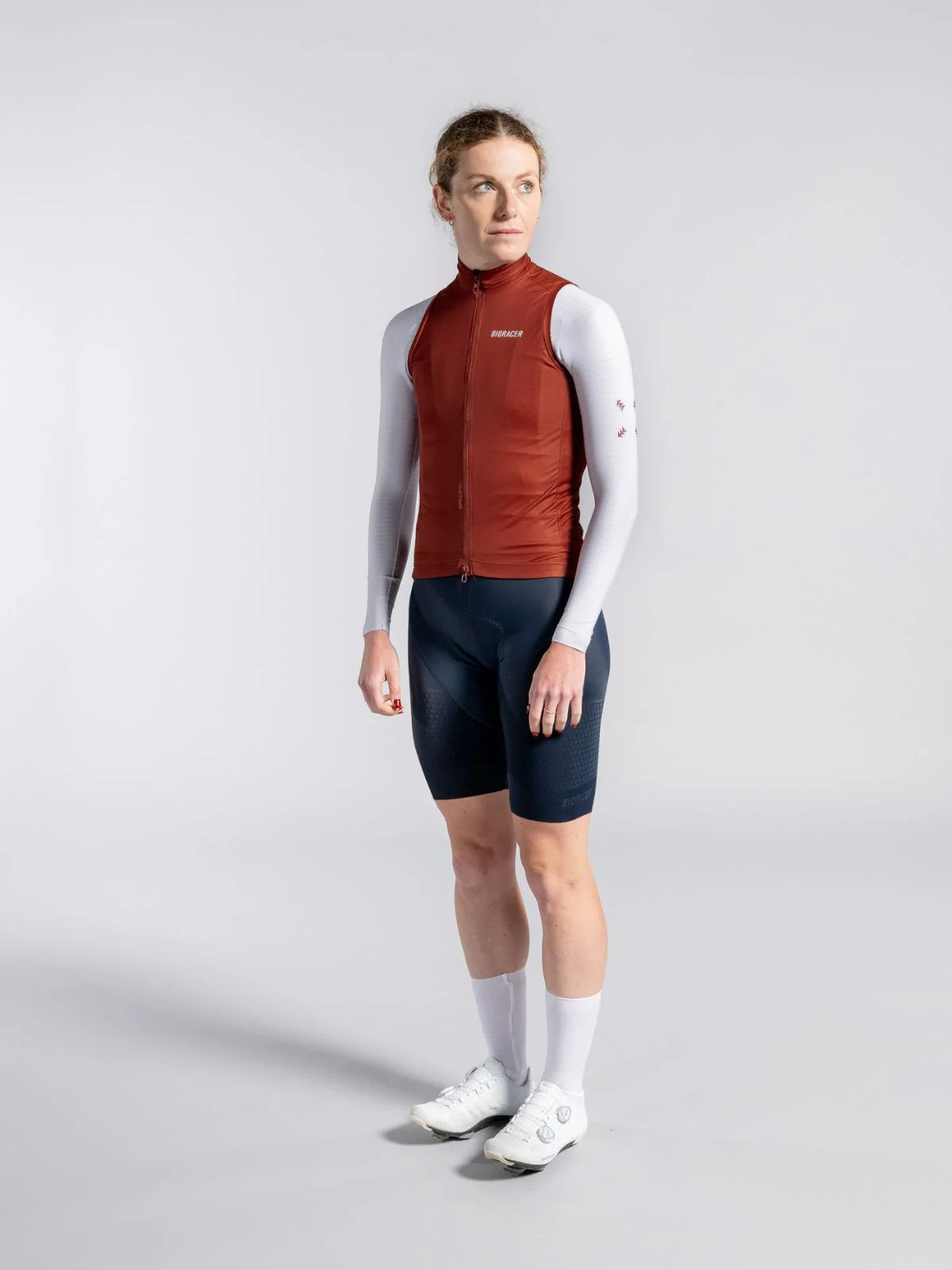 Person wearing a cycling outfit with a red vest, white long-sleeve shirt, navy shorts, and white shoes on a light gray background.